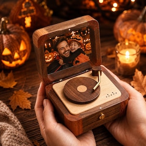 Puede incluir: Una caja de música de madera diseñada como un tocadiscos en miniatura con un marco de madera oscura y una base de madera clara. La tapa presenta una fotografía de un hombre y un niño con disfraces de Halloween. Un brazo fonocaptor dorado descansa sobre un disco de madera.