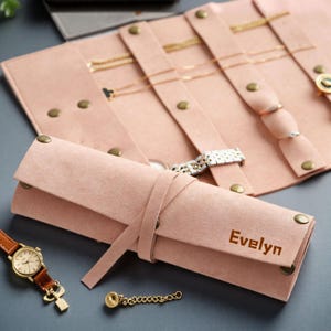 May include: A pink felt jewelry roll with the name "Evelyn" printed on the front. The roll is tied with a matching strap and holds necklaces, rings, and a watch. A gold watch with a brown leather band is also visible.