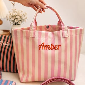 May include: A pink and white striped tote bag featuring the name Amber embroidered in orange cursive script on the front. The bag has two top handles and a single snap button closure at the center of the opening, with a glossy finish.