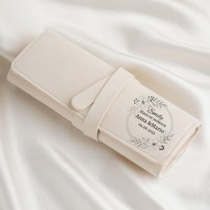 May include: A cream-colored, rolled-up accessory case with a strap closure. The case features a floral wreath design with the text "Emily Maid of Honour Anna & Mario 06.08.2025". The case is made of a soft, felt-like material.