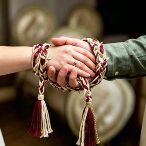 Könnte beinhalten: Ein Handfasting-Seil mit geflochtenen Strängen aus dunkelrotem Satin, cremefarbenem Seil und metallisch goldener Kordel. Das dekorative Seil ist mit kleinen goldfarbenen Blattanhängern verziert und endet an beiden Seiten in dicken, mehrfarbigen Quasten.