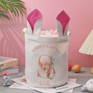 May include: A light-colored Easter basket with bunny ear handles. The basket features the text "Happy Easter" and a floral bunny illustration. The name "Sarah" is printed below the bunny. The bunny ears are pink.