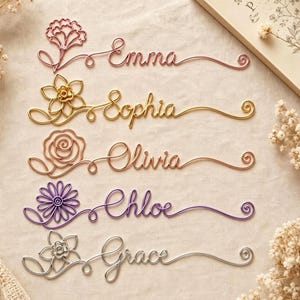 May include: Five custom wire name signs in rose gold, gold, copper, purple, and silver. Each sign features a unique wire-formed flower on the left and a cursive name: Emma, Sophia, Olivia, Chloe, and Grace, ending in a small spiral flourish.