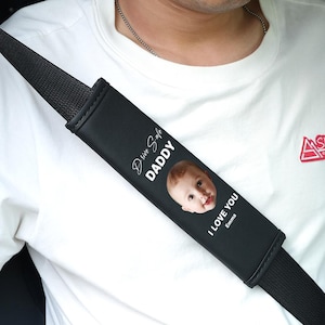 Può includere: Cuscinetto nero per cintura di sicurezza con la scritta "Drive Safe DADDY" e "I LOVE YOU Emma" stampata in bianco. Il cuscinetto presenta una foto di un bambino. La cintura di sicurezza è nera.