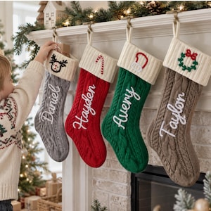 May include: Four cable-knit Christmas stockings hanging from a mantel, each featuring a cream-coloured cuff with an embroidered icon and a name in white script: Daniel with a reindeer, Hayden with a candy cane, Avery with a mitten, and Taylor with a wreath.