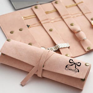 May include: A blush pink jewelry roll with a tie closure, featuring a black bow and the name "Emily." The roll is open, displaying gold necklaces, rings, and a watch. The jewelry organizer is made of soft material and has brass-colored snap closures.