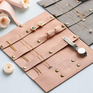 May include: A blush pink jewelry organizer with multiple compartments and brass snap closures. The organizer holds gold necklaces, earrings, rings, and a silver wristwatch. A small, white candle with a gold top is also visible.