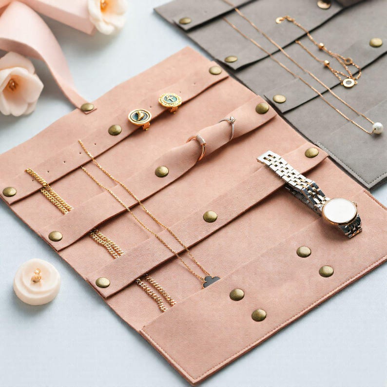 May include: A blush pink jewelry organizer with multiple compartments and brass snap closures. The organizer holds gold necklaces, earrings, rings, and a silver wristwatch. A small, white candle with a gold top is also visible.