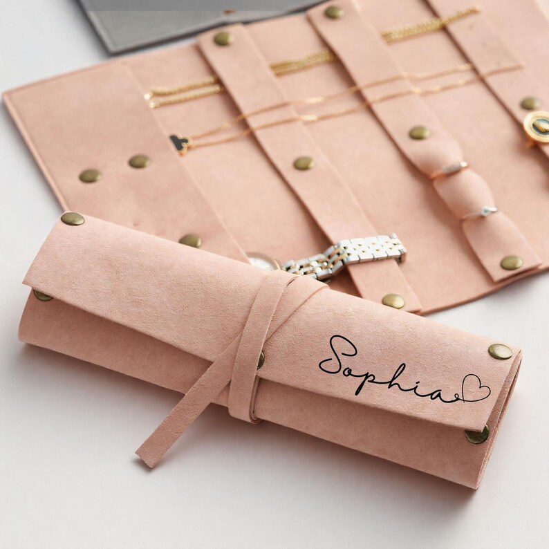 May include: A blush pink jewelry roll with a matching tie closure. The roll is adorned with the name "Sophia" in script with a heart symbol. The roll is designed to hold necklaces, rings, and a watch. The roll is secured with brass-colored snaps.