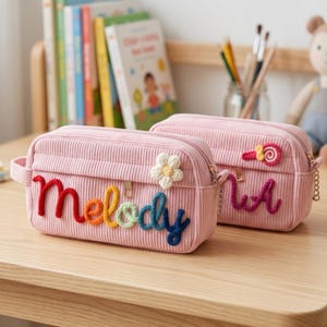 Op de afbeelding: Twee rechthoekige roze corduroy etuis met gouden ritsen. Het voorste etui heeft het woord Melody in meerkleurige garenletters en een witte gehaakte bloem. Het achterste etui toont een kleine gehaakte lolly en een deel van een woord.