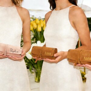 May include: Several personalized leather roll-up cases in shades of pink and brown. Each case is secured with a strap and features a name, such as "Sophia" or "Anna", in gold lettering. The cases are held in front of a background of white and yellow flowers.