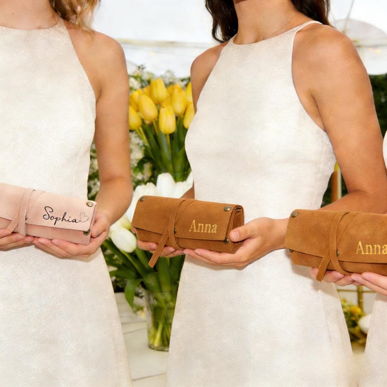 May include: Several personalized leather roll-up cases in shades of pink and brown. Each case is secured with a strap and features a name, such as "Sophia" or "Anna", in gold lettering. The cases are held in front of a background of white and yellow flowers.