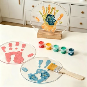 Puede incluir: Arte de huellas de manos en acrílico transparente con pintura colorida. Una pieza se exhibe sobre un soporte de madera, mostrando una huella de mano con detalles de flores azules. También se ven huellas de manos y botes de pintura adicionales.