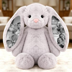 May include: A large, plush, light gray bunny rabbit stuffed animal with a pink nose and black eyes. The inside of each ear features a black and white photo collage. The bunny is sitting on a white, textured surface.