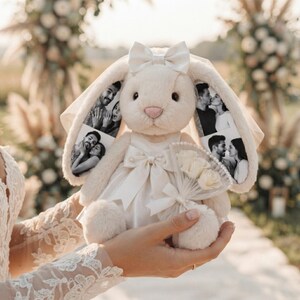 Può includere: Un coniglio di peluche color crema che indossa un abito di raso bianco con un fiocco abbinato sulla testa. Le lunghe orecchie del coniglio presentano fotografie in bianco e nero di una coppia. Tiene in mano un piccolo bouquet di fiori bianchi.