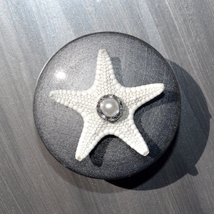 May include: A white starfish with a pearl in the center, on a black circular base.