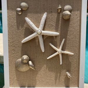 May include: A framed artwork featuring a burlap background with two white starfish and various gold-colored seashells. The starfish have a pearl-like center.