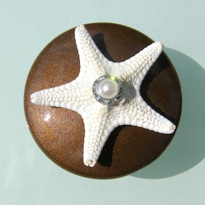 May include: A white starfish with a pearl and silver ring on a brown background.