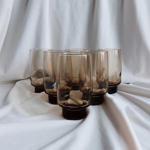 May include: Six brown-tinted drinking glasses with a dark brown base are arranged on a white cloth. The glasses have a cylindrical shape and a slightly flared rim. The base is a solid, dark brown color.