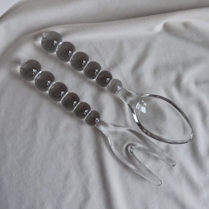May include: A clear glass salad serving set. The set includes a spoon and a fork. The handles are made of a series of clear glass beads. The serving utensils are on a white fabric background.