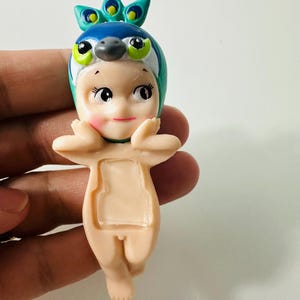 May include: A small figurine of a baby-like character wearing a peacock-themed headpiece. The figurine has a light skin tone, with a blue and green headpiece, and is holding its hands up near its face.