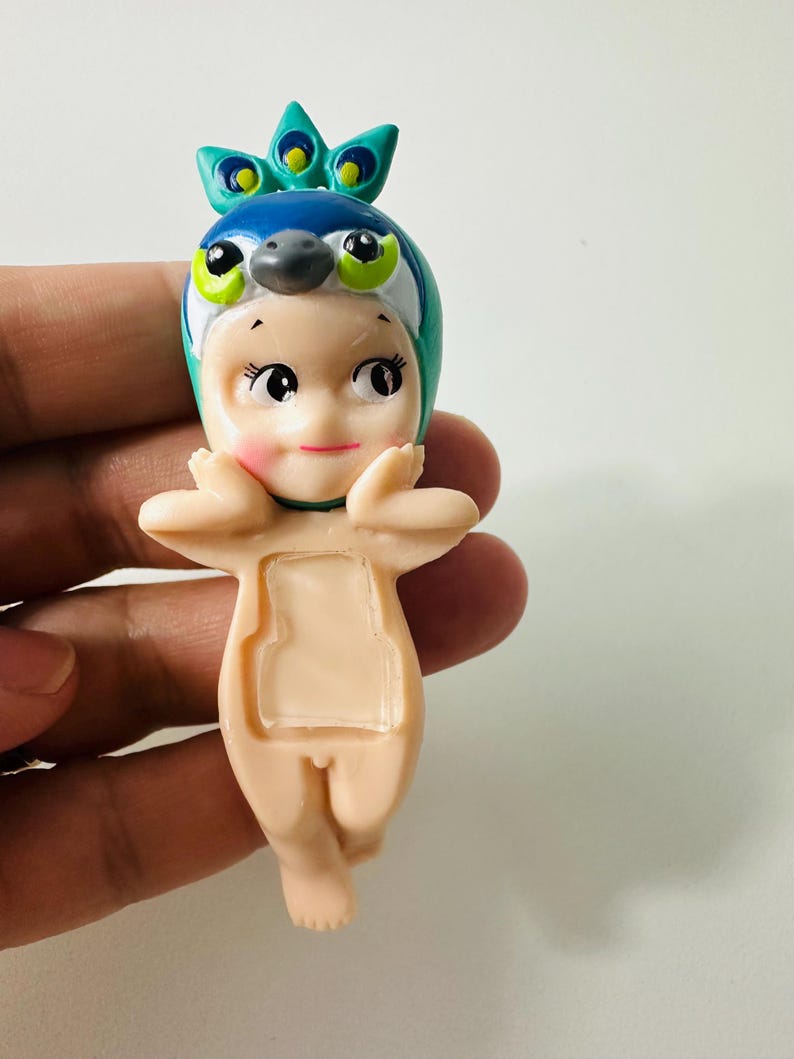 May include: A small figurine of a baby-like character wearing a peacock-themed headpiece. The figurine has a light skin tone, with a blue and green headpiece, and is holding its hands up near its face.