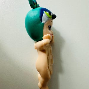 May include: A vintage figurine of a baby wearing a teal bird-themed hat with blue and white accents. The figurine has a light skin tone and is in a standing position with its hands near its face.