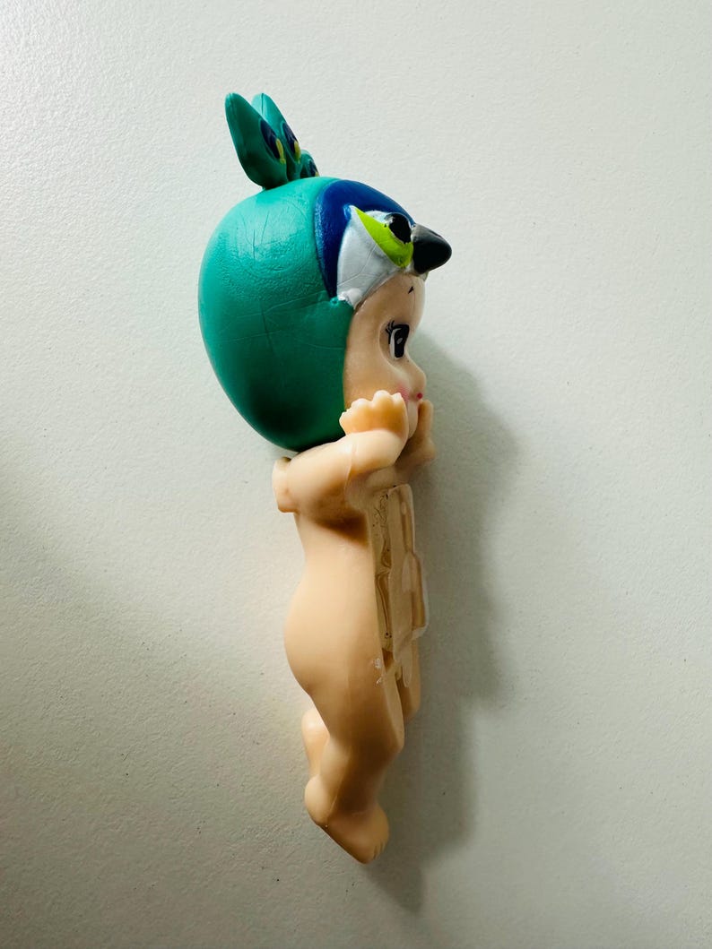 May include: A vintage figurine of a baby wearing a teal bird-themed hat with blue and white accents. The figurine has a light skin tone and is in a standing position with its hands near its face.
