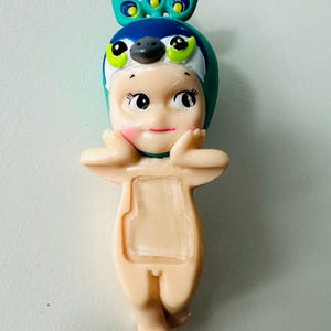 May include: A small, light-colored figurine of a baby-like figure wearing a peacock-themed hat. The hat is blue, green, and white. The figurine has rosy cheeks and is posed with its hands near its face.