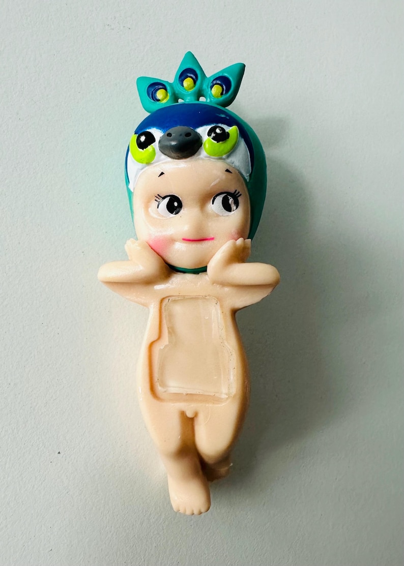 May include: A small, light-colored figurine of a baby-like figure wearing a peacock-themed hat. The hat is blue, green, and white. The figurine has rosy cheeks and is posed with its hands near its face.