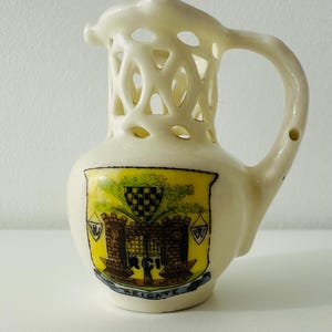 May include: Cream-colored ceramic pitcher with a decorative openwork top and a handle. The pitcher features a colorful emblem with a castle, shield, and the word "Reigate". A small, decorative piece.