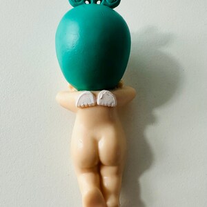 May include: A figurine of a nude cherub with small white wings, a teal-colored head, and a crown-like detail. The cherub is in a kneeling position, with its arms crossed behind its back. The figurine is made of a light beige material.