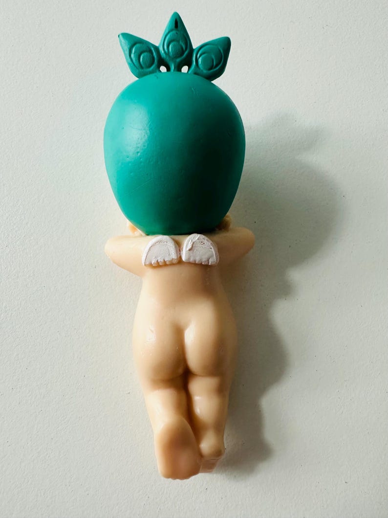May include: A figurine of a nude cherub with small white wings, a teal-colored head, and a crown-like detail. The cherub is in a kneeling position, with its arms crossed behind its back. The figurine is made of a light beige material.