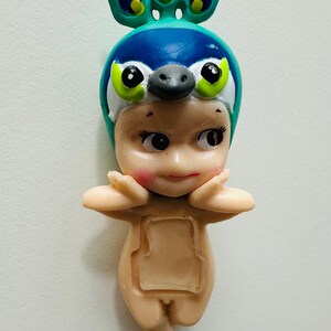 May include: A small figurine of a baby wearing a peacock-themed hat. The hat is blue and green with a beak and large eyes. The baby has rosy cheeks and is in a standing pose with arms raised.