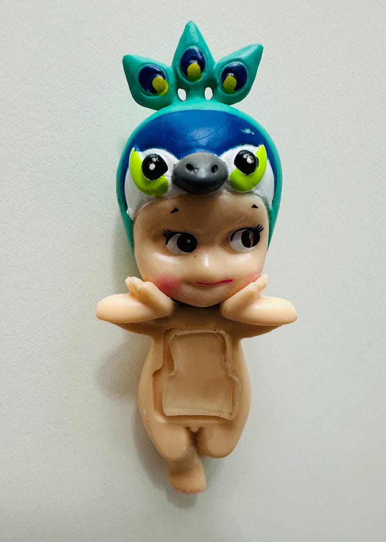May include: A small figurine of a baby wearing a peacock-themed hat. The hat is blue and green with a beak and large eyes. The baby has rosy cheeks and is in a standing pose with arms raised.