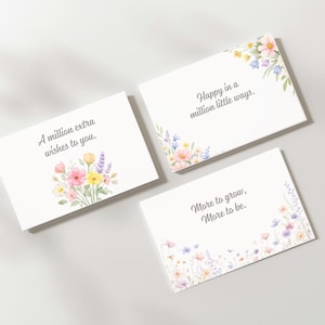 May include: Three white greeting cards with floral watercolor designs. The cards feature phrases like "A million extra wishes to you" and "Happy in a million little ways." The cards are decorated with pink, yellow, and purple flowers.