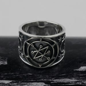May include: A wide silver-toned metal ring featuring a dark, recessed circular center with an engraved geometric sigil. The band is polished with additional small star and circular symbols etched around the outer circumference of the ring.