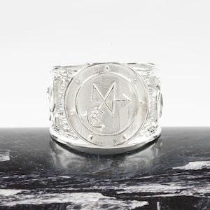 May include: A wide silver ring featuring a central circular medallion with an engraved sigil. The band has a textured, rugged finish surrounding the smooth, polished centre disc. The metal has a bright, reflective sheen throughout the entire piece.