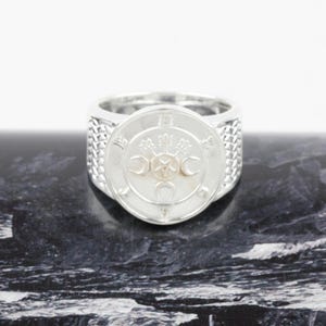 May include: A silver ring featuring a circular face with an engraved pentagram surrounded by three crescent moons. The band has a textured, cross-hatched pattern on the sides. The text BHALA JEWELLERS is visible in a small black logo in the corner.