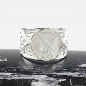 May include: A wide silver ring featuring a circular centerpiece engraved with the Sigil of Lucifer. The band has a textured, hammered finish on the sides and a polished, smooth surface on the circular emblem. The logo for Bhala Jewellers is visible.