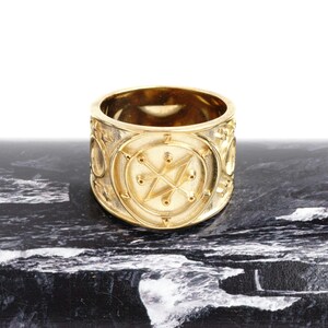 May include: A wide gold band ring featuring a central circular emblem with a raised star-like geometric design and small dot accents. The polished metal surface reflects light, highlighting the intricate engraved details on the exterior circumference.
