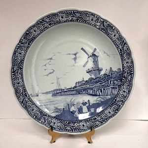 May include: A large, round, blue and white decorative plate. The plate features a detailed landscape scene with a windmill, buildings, and figures near a body of water. The rim has a scalloped edge and a floral and scroll design.