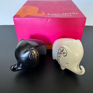 May include: A black and white elephant-shaped salt and pepper shaker set. The black shaker has gold accents, while the white shaker has black accents. The set comes with a pink box with the words "kate spade NEW YORK".