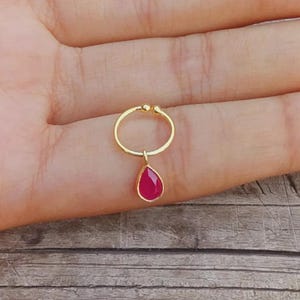 Peut inclure: Un anneau de nez doré en forme de cercle ouvert avec deux petites extrémités arrondies, agrémenté d'une breloque en forme de goutte sertie d'une pierre précieuse rose vif. La pierre est entourée d'un fin contour en métal doré.