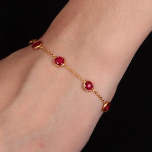 May include: A gold-colored bracelet featuring a delicate chain design. The bracelet is adorned with several round, vibrant red gemstones. The gemstones are evenly spaced along the chain, creating a simple yet elegant look. The bracelet is shown against a dark background.