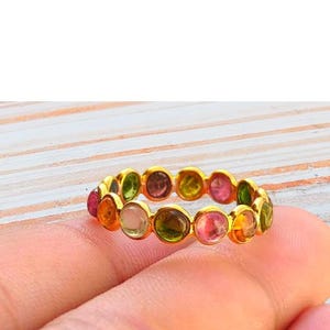 May include: A gold band ring featuring a continuous row of small, round, bezel-set multi-colored tourmaline gemstones. The stones display a variety of hues including pink, green, yellow, and clear tones, all set within a polished gold metal frame.