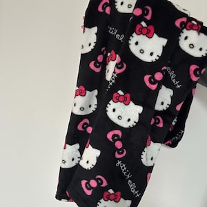 Peut inclure: Couverture en peluche noire avec un motif répété de visages Hello Kitty blancs avec des nœuds roses et des accents de nœuds roses. La couverture porte également le texte "fitti ellott" en écriture blanche.