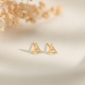 May include: A pair of gold-colored stud earrings, each shaped as the letter "A". The earrings are embellished with small, clear gemstones. The earrings are displayed on a white surface, with a blurred floral arrangement in the background.