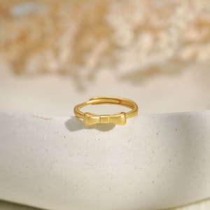 May include: A gold-toned ring featuring a small, centered bow detail. The band is thin and circular, with a smooth, matte finish. The bow is positioned at the top of the ring, showcasing a simple, clean design with no additional stones or engravings.