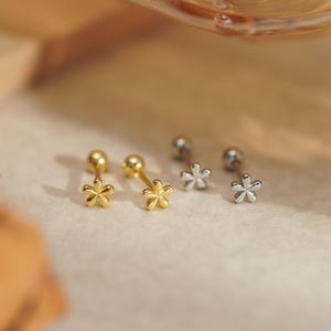 May include: Four flower stud earrings, two gold and two silver. Each earring features a small ball and a flower design. The earrings are presented on a light-coloured surface. The earrings are small, suitable for everyday wear.